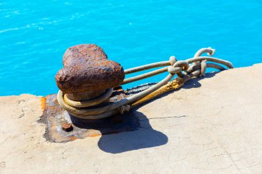 Paslı çapa iskelede bir ipe bağlı. Yıpranmış çapa iskeledeki beton duvara monte edilmiş.