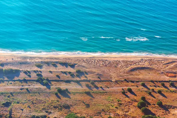 stock image View from above a sandy beach with a coastal road in the background. Beach is lined with trees and bushes, creating a serene and peaceful atmosphere