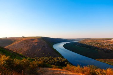 Nehir, üzerinde açık mavi bir gökyüzü olan bir vadiden akar. Sahne huzurlu ve sakin, su gökyüzünü ve çevreyi yansıtıyor. Tepeden manzara nehir manzarası