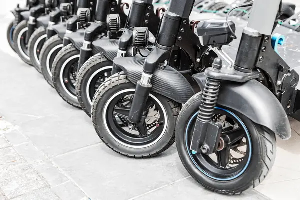 Scooter 'lar şehrin kaldırımında arka arkaya park etmiş. Sokakta dizilmiş siyah kiralık elektrikli scooterlar.