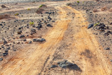 Arka planda toprak ve taşlı toprak yol. Yol asfalt değil ve oldukça pürüzlü. Çorak bir çöl manzarasında kestirme yol.