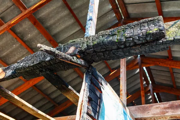 stock image Charred log is hanging from the ceiling of a building. Charred wood has a dark, smoky appearance and is surrounded by a grey roof. Concept of destruction and decay, as the wood has been burned