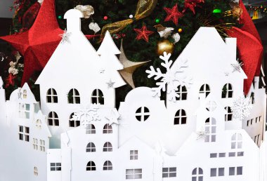 Güzel dekore edilmiş bir Noel ağacının altına yerleştirilmiş beyaz kağıt kesiği bayram manzarası yaratıyor. Üzerinde evler olan Noel ağacı.