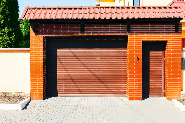 Kırmızı fayanslı çatı altında dış garaj kapısı, klasik mimari stil ve canlı renk kontrastı sergiliyor. Tuğla duvarlı ve kahverengi kapılı bir garaj.