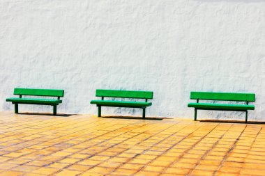 Yeşil banklar beyaz bir duvarın önündeki kaldırımda oturuyorlar. Ahşap banklar boş ve bölge sessiz.