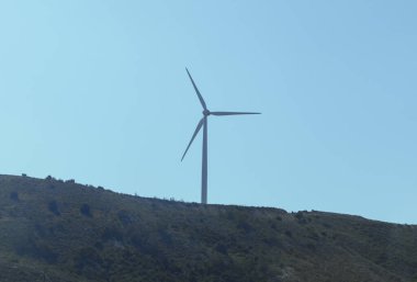 Rüzgar türbini açık mavi gökyüzü olan bir tepede. Rüzgar türbini yeşil bir yamaçta dimdik durur, açık mavi gökyüzüne karşı yenilenebilir enerji sağlar.