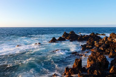 Okyanus kayaları kıyı şeridi boyunca yayılmış. Derin mavi sular ve yumuşak dalgalarla çevrili engebeli kayalar.