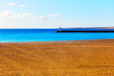 Sahil sahnesinde hafif dalgalar, açık bir gökyüzüne karşı dik duran deniz feneri.