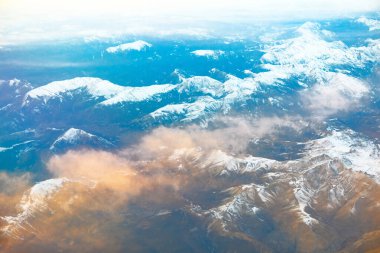 Güneş doğarken karla kaplı dağ tepelerinin havadan görünüşü kısmen bulutlar tarafından gizleniyor. Engebeli arazi, doğal güzelliği ve manzaranın ihtişamını vurgulayan yumuşak bulutlarla çelişiyor..