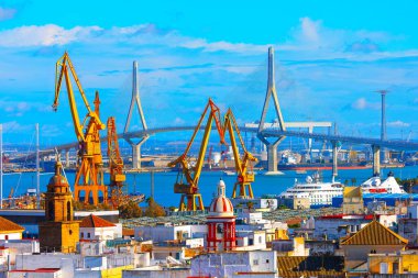 Cadiz İspanya, sarı vinçleri, asma köprüsü ve tarihi ve modern binaların karışımına sahip canlı bir liman kenti sergiliyor. Sahne berrak gökyüzü ve liman faaliyetleriyle dolu.