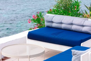 Açık hava oturma alanı sakin, mavi bir denize bakıyor. Mavi ve gri yastıklı beyaz çerçeveli bir kanepe yuvarlak beyaz bir masanın yanında, pembe çiçekli yeşil bitkilerle çevrili, dingin ve rahatlatıcı bir yer yaratıyor.