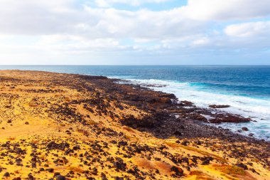 Kanarya Adaları 'ndaki kayalık bir kıyı şeridi, kumlu, kurak bir arazi ve karanlık kayalara çarpan okyanus dalgalarıyla kıyı manzarası. Parçalı bulutlu gökyüzü canlı, dinamik okyanusla çelişiyor, doğal güzelliği ve sertliği vurguluyor.