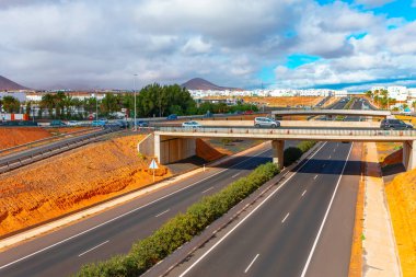 Highway in Arrecife, Lanzarote, with an overpass bridge crossing over it. Highway is divided by a median strip with greenery, there are multiple lanes in each direction. Overpass road with vehicles  clipart