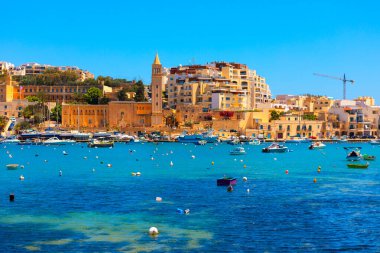 Malta 'nın sahil kenti Marsaskala, küçük tekneleri ve şamandıraları olan canlı mavi sulara sahip. Ufuk çizgisi geleneksel ve modern mimari ile harmanlanır ve bir kilise kulesi içerir.