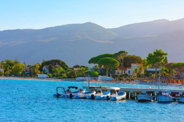 Palermo, Sicilya 'daki Mondello Plajı. Bir rıhtıma kenetlenmiş birkaç teknenin bulunduğu kıyı sahnesi yemyeşil ve dağlık bir zemin üzerine kurulmuş. Deniz suyu mavi ve bölge huzurlu görünüyor.