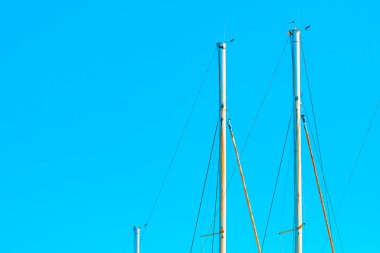 Yelkenlinin üst kısımları açık mavi gökyüzüne karşı. Direkler, tepelerinde çeşitli donanım hatları ve küçük cihazlar ile donatılmıştır, canlı gökyüzü kontrastı nedeniyle görsel bir sahne oluşturur