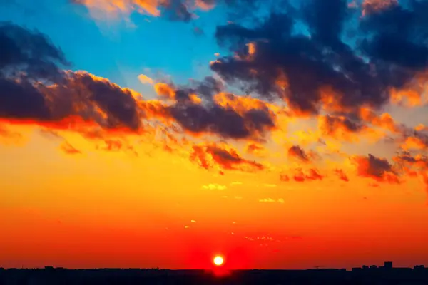 Güneş ufukta batarken, gün batımı canlı, gökyüzünde parıldıyor. Kara bulutlar gün batımının parlak renkleriyle dramatik bir tezat oluşturuyor. Alacakaranlıktaki uzak şehir manzarasının silueti