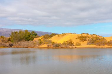 Ön planda göl olan sakin bir manzara, kumlu kumullar ve arka planda seyrek bitki örtüsü. Gökyüzü çoğunlukla bulutlu, mavi lekeler var ve bazı uzak dağlar görülebilir.. 