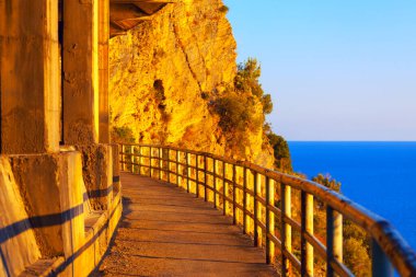 Altın günbatımı ışığıyla aydınlatılan metal parmaklıklarla kaplı eğimli uçurum kenarı yolu, parlak turuncu kaya yüzeyine ve berrak bir gökyüzünün altında derin mavi denize karşı tezat oluşturuyor. Bir uçurum boyunca yürüyorum
