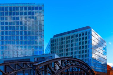 Hamburg 'daki demir köprü yapısının arkasındaki mavi renkli cam cepheli modern ofis binaları. Sahne, Hamburg gibi kentsel manzaralarda bulunan modernlik ve tarihsel unsurların karışımı.