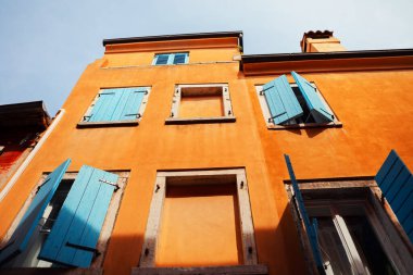 Mavi ahşap panjurlu parlak renkli turuncu bina cephesi, bazıları açık, bazıları kapalı. Canlı turuncu duvarlar ve mavi panjurlar arasındaki zıtlık estetik açıdan memnun edici bir manzara yaratır.