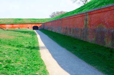 Yeşil çimenler ve çimenli bir set ile çevrili kemerli bir kapısı olan tuğla bir duvara giden yol. Tuğla duvar ve kemerli tarihi kaleler eski bir kaleye giriş olduğunu gösteriyor. 