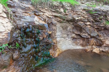 Kayalık bir uçurumdan aşağıdaki su havuzuna akan küçük bir şelale. Kayalardaki doğal erozyon şekilleri ve yeşil yosun veya yosunların varlığı nemli bir çevreyi işaret ediyor.