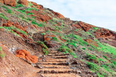 Yeşil bitki örtüsü ve kırmızımsı toprakla kaplı bir yamaçtan yükselen taş basamaklardan oluşan engebeli bir açık hava merdiveni. Doğal güzellik ve dik araziyi yürüyüşçüler için ulaşılabilir kılma çabası