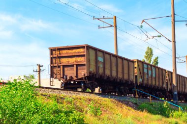 Yükseltilmiş raylarda giden vagonlu bir yük treni. Elektrik hatları ve direkler üzerinde, demiryolu yakınında yeşillik ve arka planda açık mavi bir gökyüzü ile