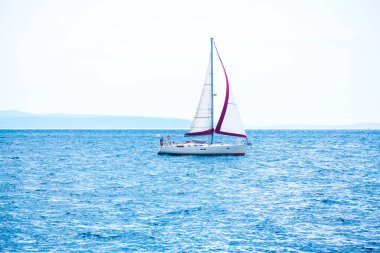 Yelkenli, berrak mavi gökyüzünün altında sakin okyanus sularında süzülüyor. Beyaz yelkenli bir yat engin mavi sularda yüzer. Uzak diyarların belli belirsiz dış hatlarıyla sakin bir deniz manzarası yaratıyor.
