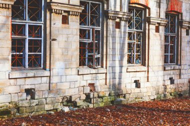 Hasarlı duvarları, parmaklıklı camları ve zemini kaplayan düşmüş yaprakları olan eski bir taş binanın yakın çekimi. Yaşlanmış mimari ve doğal çürüme tarih, nostalji ve terk edilme duygusunu çağrıştırıyor.