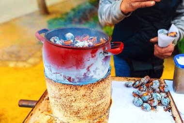 Bir sokak satıcısı, taşra tabanında kırmızı demlik kullanarak geçici bir sobanın üzerinde kestane pişiriyor. Dumanlı aroma taze kavrulmuş kestanelerin etrafını sararken, kağıt külahları istekli müşteriler için servis yapıyor.