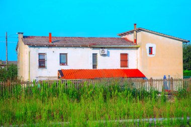 Döşeme çatılı, parmaklıklı pencereli kır evi. Etrafı fazla gelişmiş bitki örtüsü ve ahşap çitlerle çevrili.