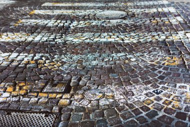 Dairesel desenli, rögar kapaklı ve kanalizasyon ızgaralı bir kaldırım taşı caddesi. Çakıl taşları beyaz ve sarı renklerle işaretlenmiş. Trafik veya yaya yönergelerini gösteriyor.     