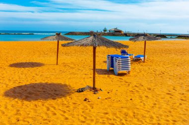 Kuma gölge düşüren saman şemsiyeli plaj sahnesi ve mavi-beyaz şezlonglar. Sakin ve dalgalı okyanuslarla dolu bir arka plan sakin ve davetkar bir deniz kenarı konumu öneriyor.