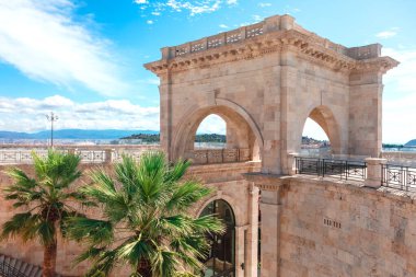 Bastione di Saint Remy, Cagliari, Sardinya İtalya 'da. İki büyük kemerli tarihi taş kemer yapısı, palmiye ağaçları ve arka planda deniz ve dağlar manzarası ile çevrili.