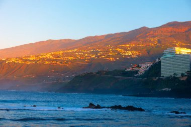 Tenerife 'deki Puerto de la Cruz sahili, kayalık oluşumları ve arka planda dağlık manzaralı okyanus manzarası. Otelli sahil şeridi ve batan güneş bölgeye sıcak bir ışık saçıyor.