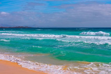 Turkuaz dalgaların kumlu sahile çarptığı sahilin manzarası. Parçalı bulutlu gökyüzünün altında ufukta görünen uzak dağların altında. Sahilde zümrüt yeşili dalgalarla sakin bir sahil manzarası