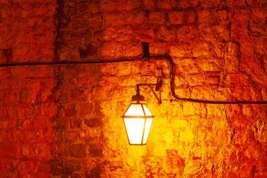 Single lamp illuminates an old stone wall, casting warm glow. Warmly lit, vintage style lantern mounted on textured stone wall, visible electrical wiring running horizontally and vertically along wall clipart