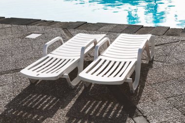 İki beyaz şezlong havuz kenarında yan yana oturur, güneş ışığında gölgeler bırakır. Sakin mavi su gökyüzünü yansıtıyor rahatlatıcı ve davetkar atmosfer yaratıyor tatil ve eğlence teması için mükemmel
