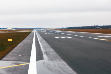 Bulutlu bir gökyüzünün altında, uzun ve boş bir havaalanı pisti. Pistin her iki tarafında da çimenlik alanlar olan beyaz çizgiler ve numaralar var.