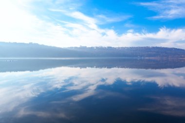 Sakin göl, gökyüzünün berrak bir yansıması ve sakin yüzeyinde uzak tepeler. Gökyüzü ince bulutlarla parçalı bulutludur ve ufukta tepeler üzerinde binalar ya da yapılar bulunur.