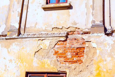 Eski ve yıkıcı bir binanın dış cephesinde gözle görülür bir hasar var. Alçı birkaç yere düşmüş, altta yatan kırmızı tuğla ve duvarın tepesine yakın dekoratif elementler var.