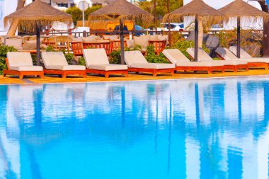 Havuz kenarında bej şezlonglu sandalyeler mavi suya yansıyan şemsiyelerin altında sıralanmış. Ayarlar dinlenme ve rahatlık için tasarlanmış, dinlenme tesisinin açık hava alanı gibi görünüyor