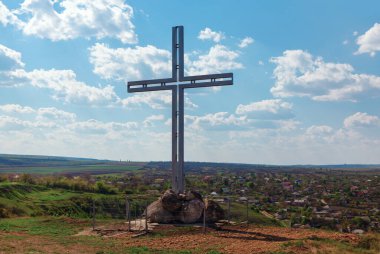 Tepenin üstünde duran büyük metal haç, sakin bir köye ve bulutlu bir gökyüzünün altındaki geniş alanlara bakıyor. Kayaların tabanında haç var ve etrafı çitlerle çevrili, önemli bir yer.