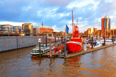 Hamburg rıhtım sahnesi, diğer gemilerin yanında demirli kırmızı gemi, modern mimari ve dramatik bir gökyüzü mükemmel bir zemin oluşturuyor. Hamburg Almanya 'daki liman sahnesinde çeşitli tekneler kenetlendi