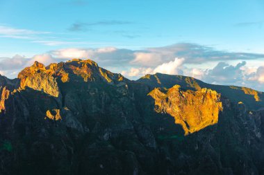 Madeira 'nın nefes kesici manzarası, altın güneş ışığıyla yıkanmış engebeli tepeler, dramatik gölgeler arazinin dokusunu güçlendiriyor. Bulutlu gökyüzü çarpıcı bir kontrastı ekliyor, adanın doğal güzelliğini vurguluyor 
