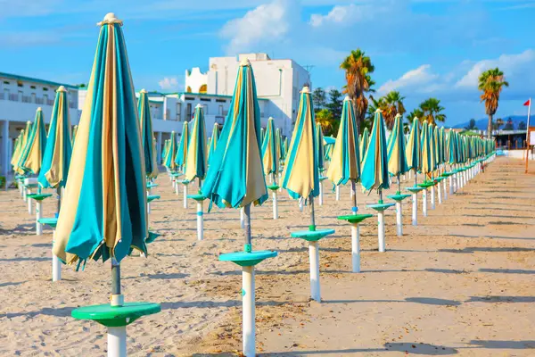 Kapalı, renkli şemsiyeli bir kumsal. Bina ve palmiye ağaçlarının arka planı mavi gökyüzünün altında, dağınık bulutlarla durmakta ve sakin sahil atmosferine katkı sağlamaktadır.