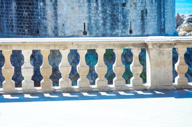 Karmaşık oyulmuş balyalarla dolu taş balyaları ön planda duruyor, fayanslı yüzeyde gölgeler bırakıyor. Arkasındaki yıpranmış taş duvar ve denizin bir bakışı tarihi bir kıyı bölgesini işaret ediyor.