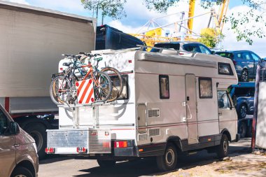 Arka rafta bisikletleri olan beyaz bir karavan otoyolun yakınına park edilmiş, diğer araçlar ve bir inşaat vinci ile çevrili. Manzara gelişmekte olan bölgede bir dinlenme alanı olduğunu gösteriyor